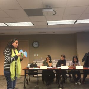 Racialized woman holding cards, speaking to a table of other racialized women