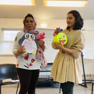 Two racialized women holding up craft items of a penguin and an emoji face