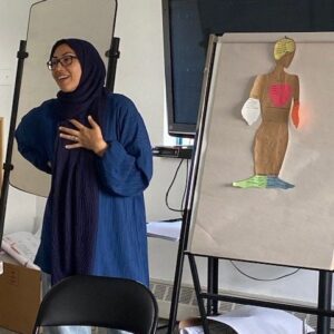 racilaized woman with blue headscarf presenting in front of an art image of a person